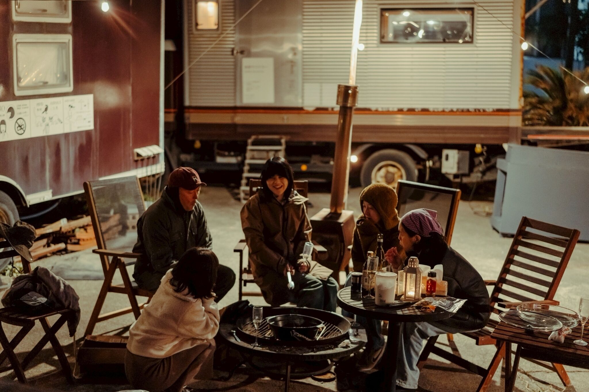 A group of people seated around a table with a portable grill, in a setting with a trailer and a vehicle in the background.