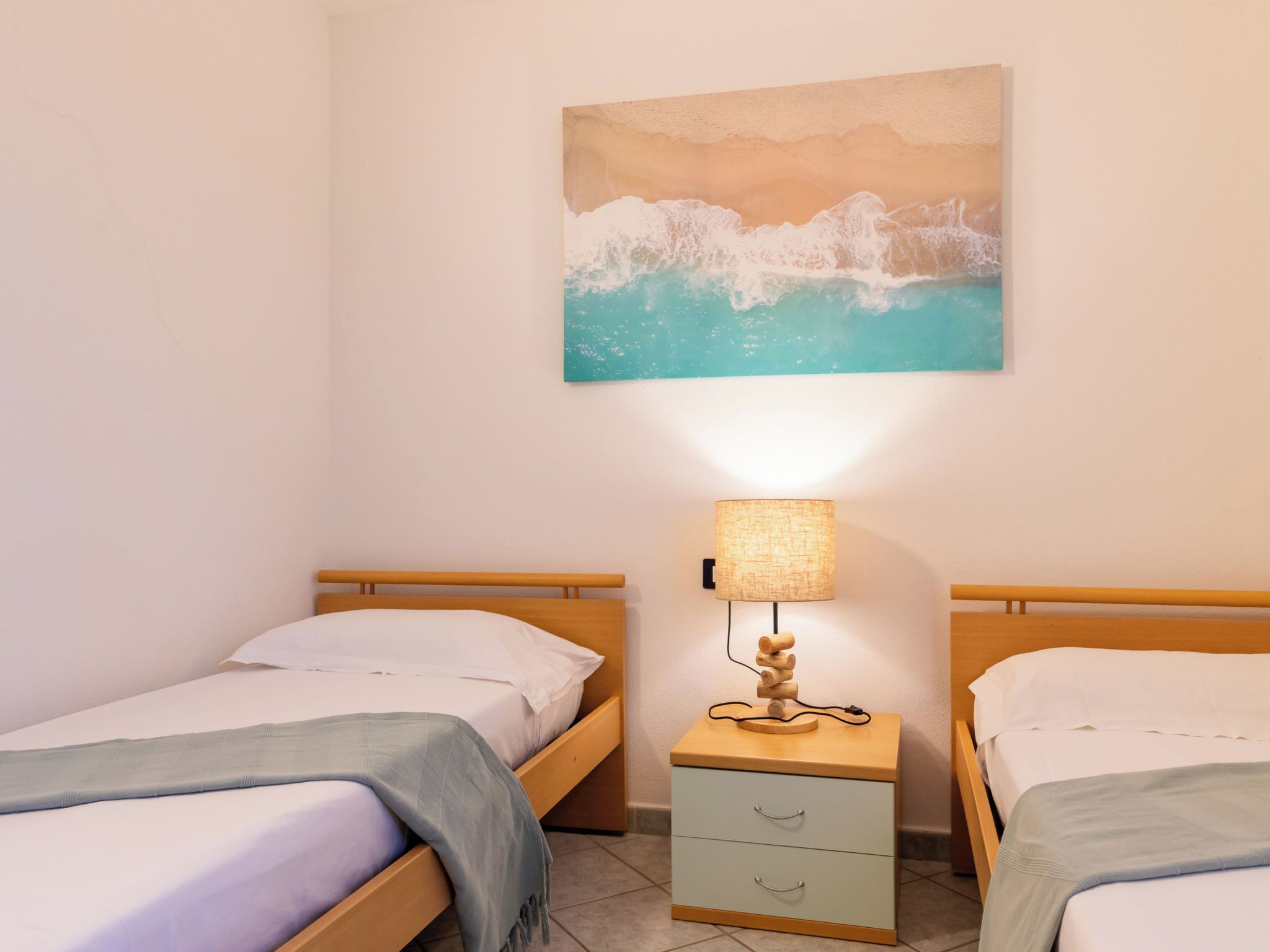 Una habitación con dos camas, una mesita de noche con lámpara y un cuadro de una playa.