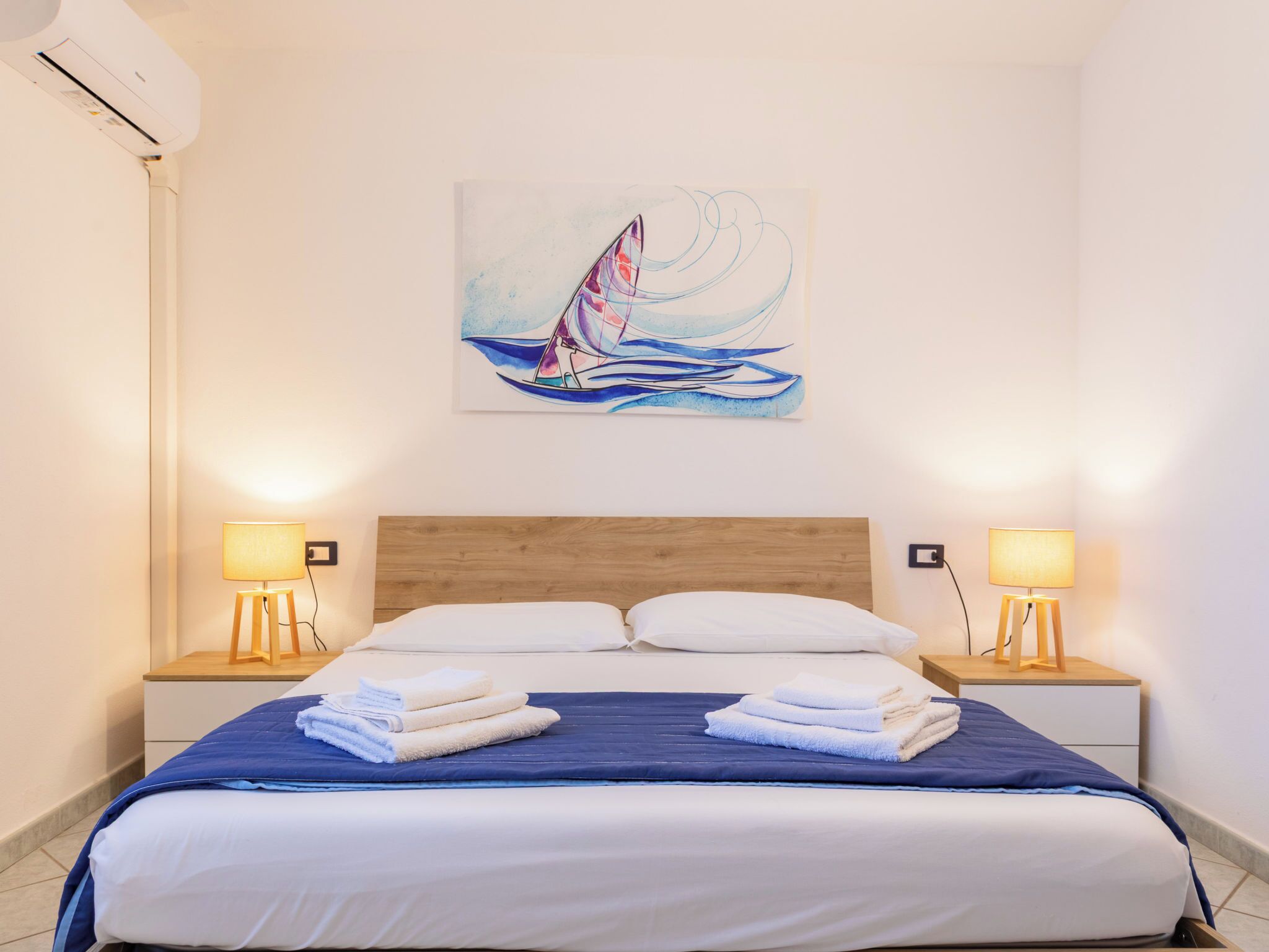 A hotel room with a wooden headboard, a bed with blue and white bedding, two bedside lamps, and a sailboat painting on the wall.