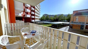 A balcony with white plastic chairs and a table, overlooking a courtyard with buildings and trees.
