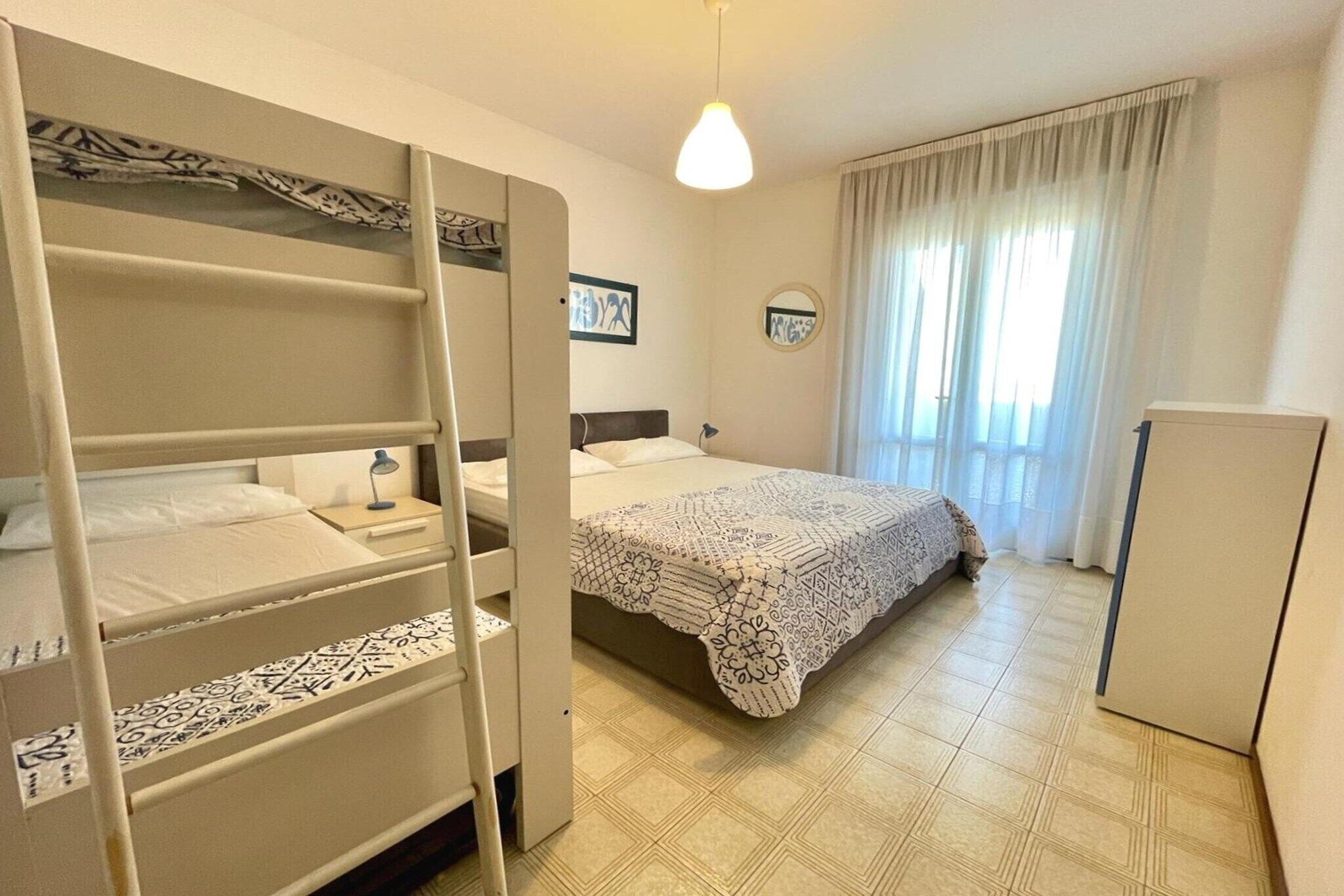 Une chambre avec des lits superposés, un lit simple, une table de chevet avec une lampe, une armoire et une fenêtre avec des rideaux.