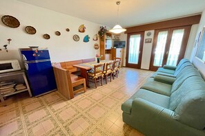 A living room with a sofa, dining table, chairs, a blue refrigerator, and a kitchen area with cabinets and a microwave.