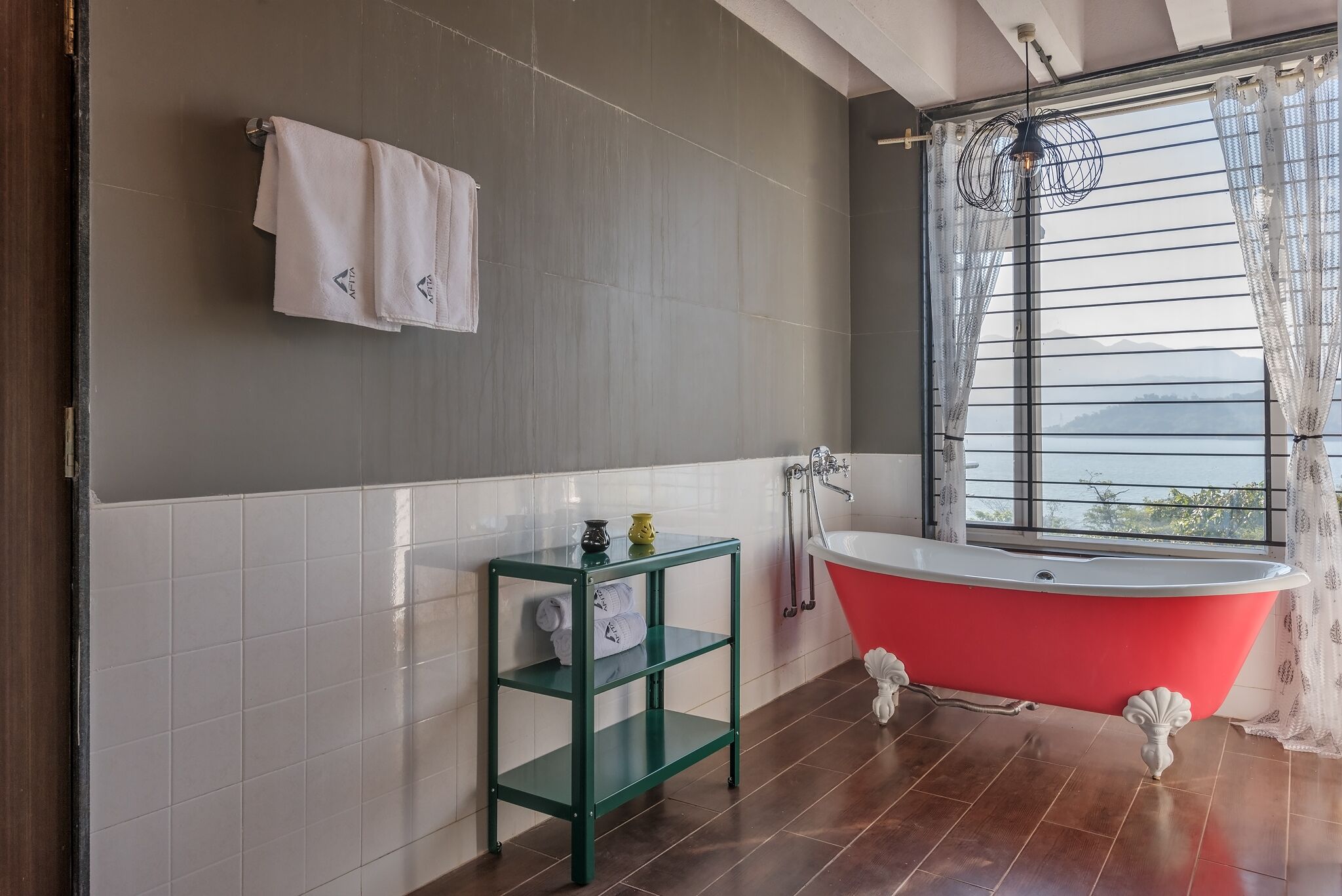 Un baño con una bañera roja estilo pata de león, un estante verde con artículos de tocador, una toalla blanca colgada en la pared y una ventana con vistas a las montañas.