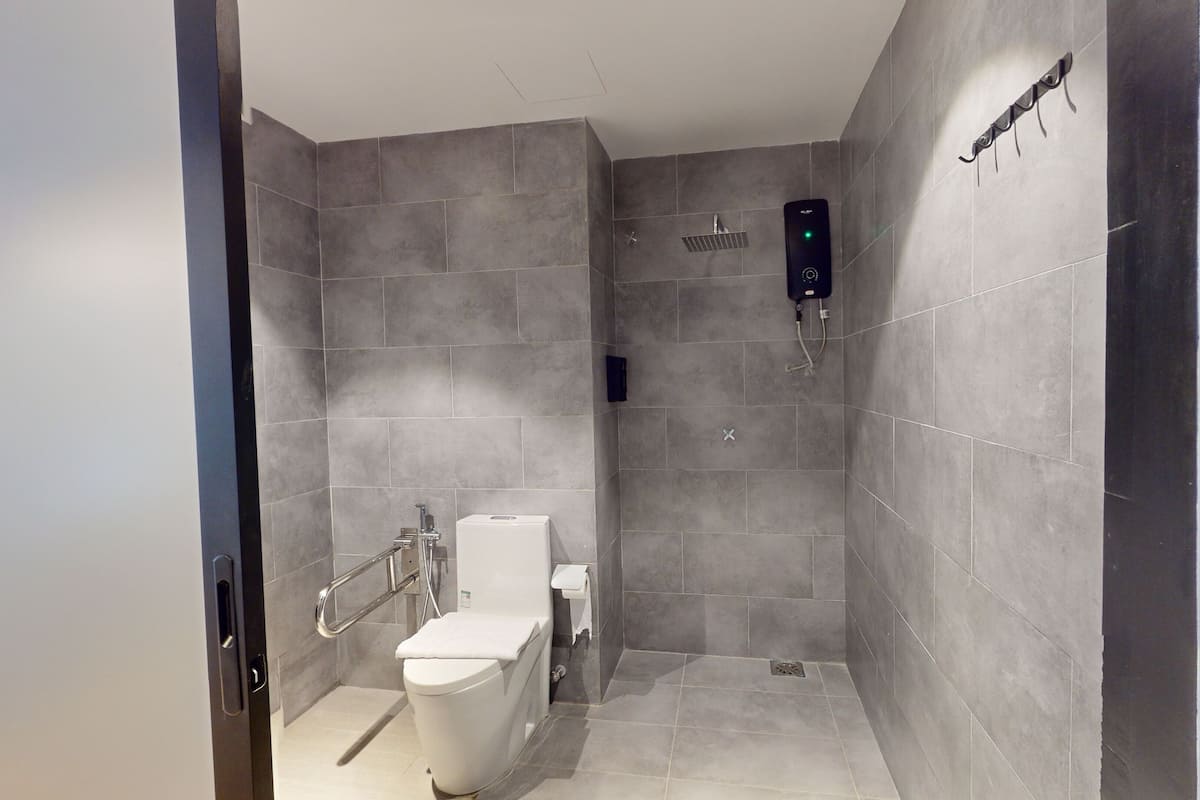 A modern bathroom with grey tiles, a white toilet, a shower with a control panel, and a handrail.