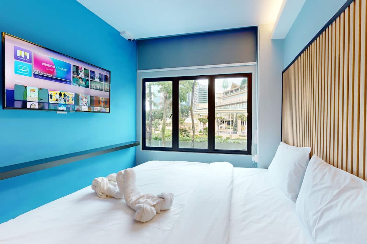 A modern hotel room with a large bed, a window with a view of a building and greenery, and a wall-mounted TV displaying various channels.