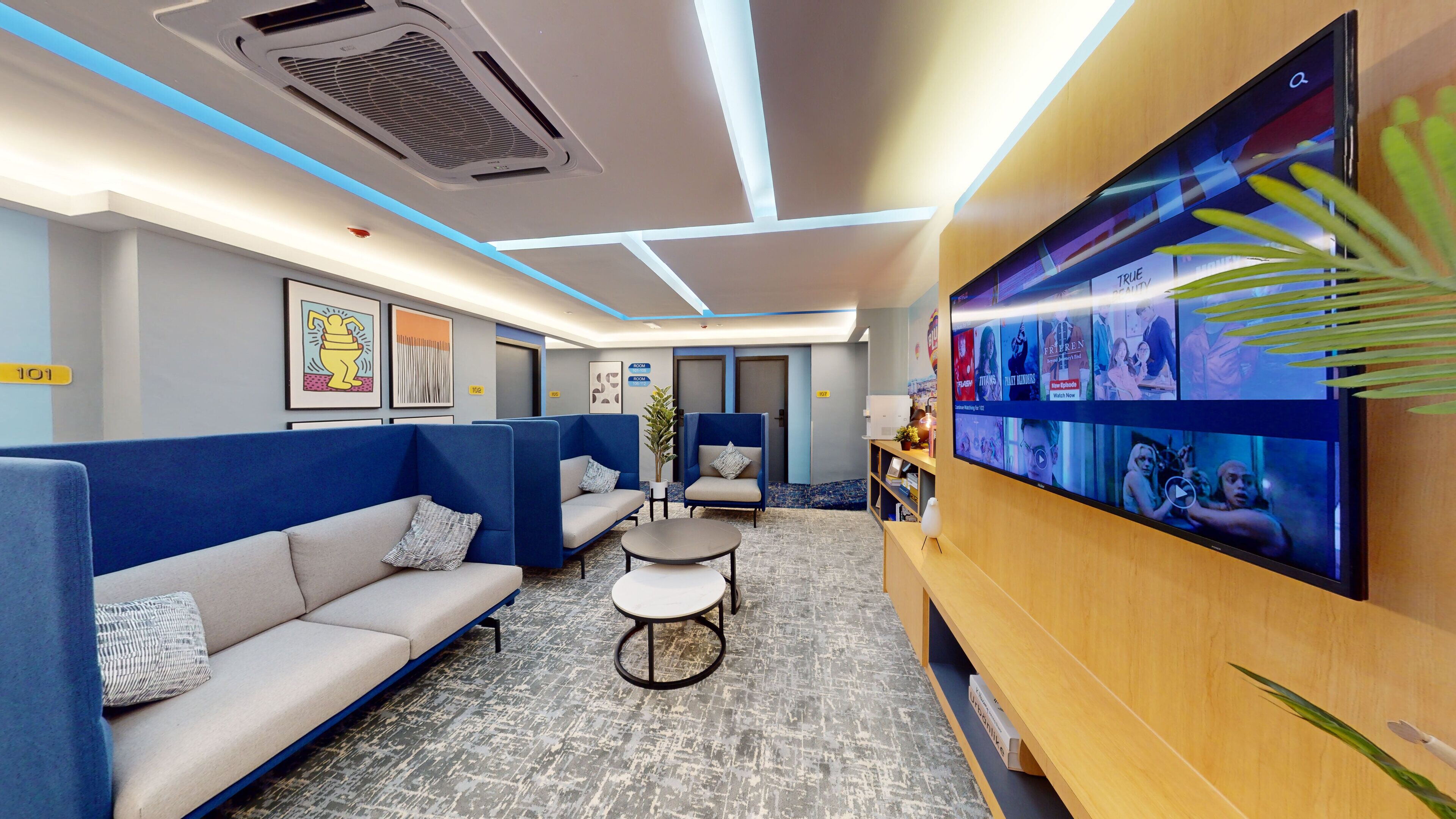 A modern waiting area with blue and grey seating, a flat-screen TV displaying various shows, and a ceiling with recessed lighting.