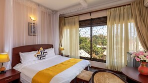 A hotel room with a large bed, a desk with a lamp, a chair, and a window with curtains. - ENTEBBE TRAVELLER'S HOTEL (Entebbe)