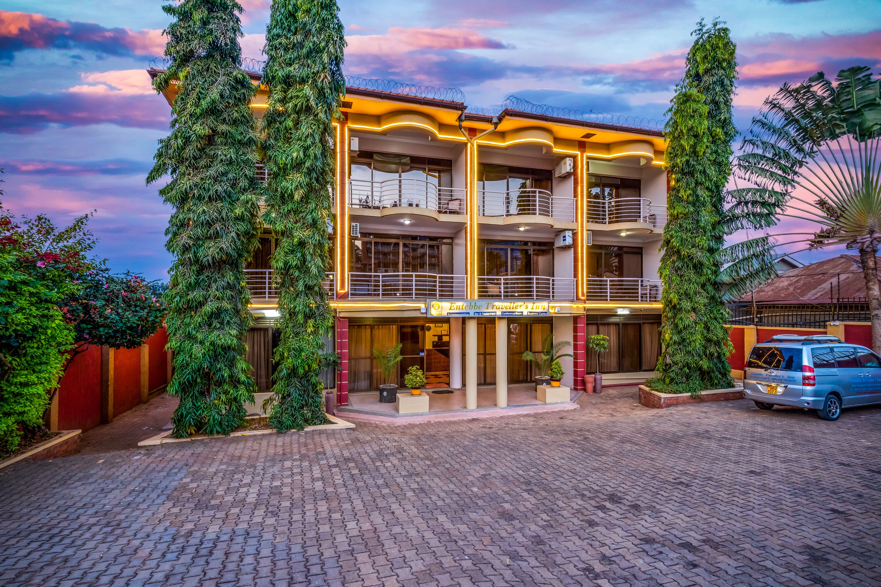 A multi-story building with balconies, a sign for 'Entebbe Traveller's Inn,' and a parked van.