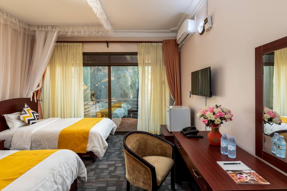 A hotel room with two beds, a desk, a chair, a TV, a mirror, and a window with curtains.
