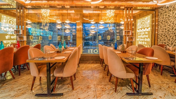 Restaurante com teto de madeira, luminárias pendentes e vista através de grandes janelas. Mesas montadas com pratos e copos.
