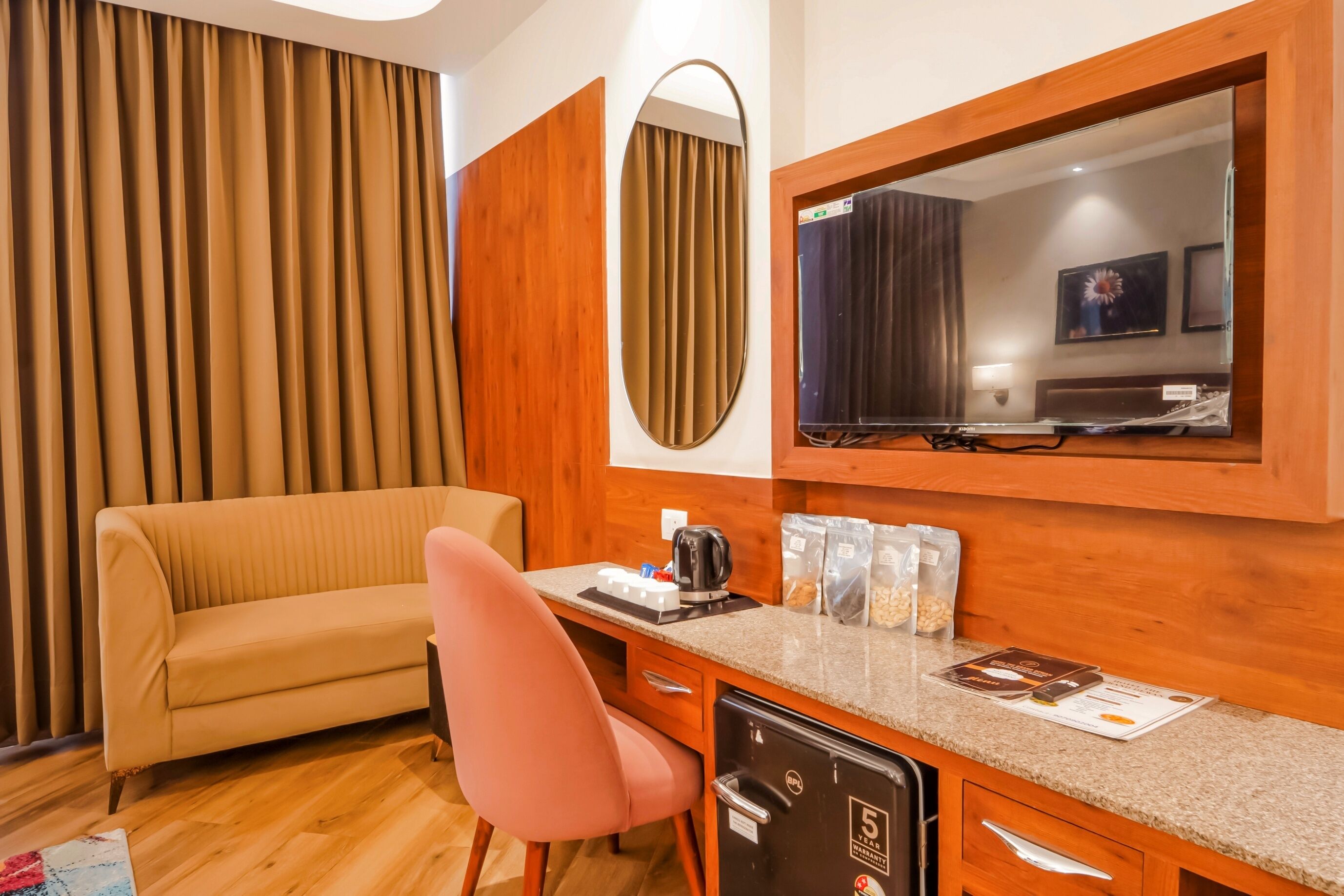 A hotel room with a beige sofa, a pink chair, a desk with a kettle, a mini-fridge, and a large mirror.