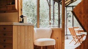 Een knus houten interieur met een zachte fauteuil, een houten kast met lades, en uitzicht op groen buiten door grote ramen.