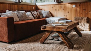 Een knus ingerichte kamer met een houten bank, een houten bijzettafeltje met een boek en een decoratieobject, plus een geweven vloerkleed.