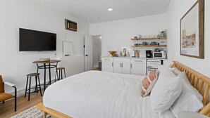 Une chambre avec un grand lit, un bureau avec deux chaises, une télévision, un coin cuisine avec des placards et des appareils électroménagers, et un tableau encadré au mur.