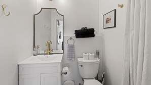 Une salle de bain avec un lavabo blanc, un robinet doré et un miroir au-dessus. On y trouve des toilettes avec deux rouleaux de papier toilette et une serviette noire accrochée au mur.
