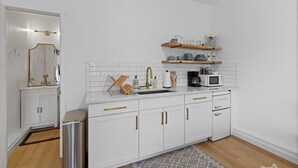 Une cuisine moderne avec des meubles blancs, un évier en acier inoxydable et un four à micro-ondes. Des étagères en bois sont fixées au-dessus du plan de travail et servent à ranger la vaisselle et une petite plante.
