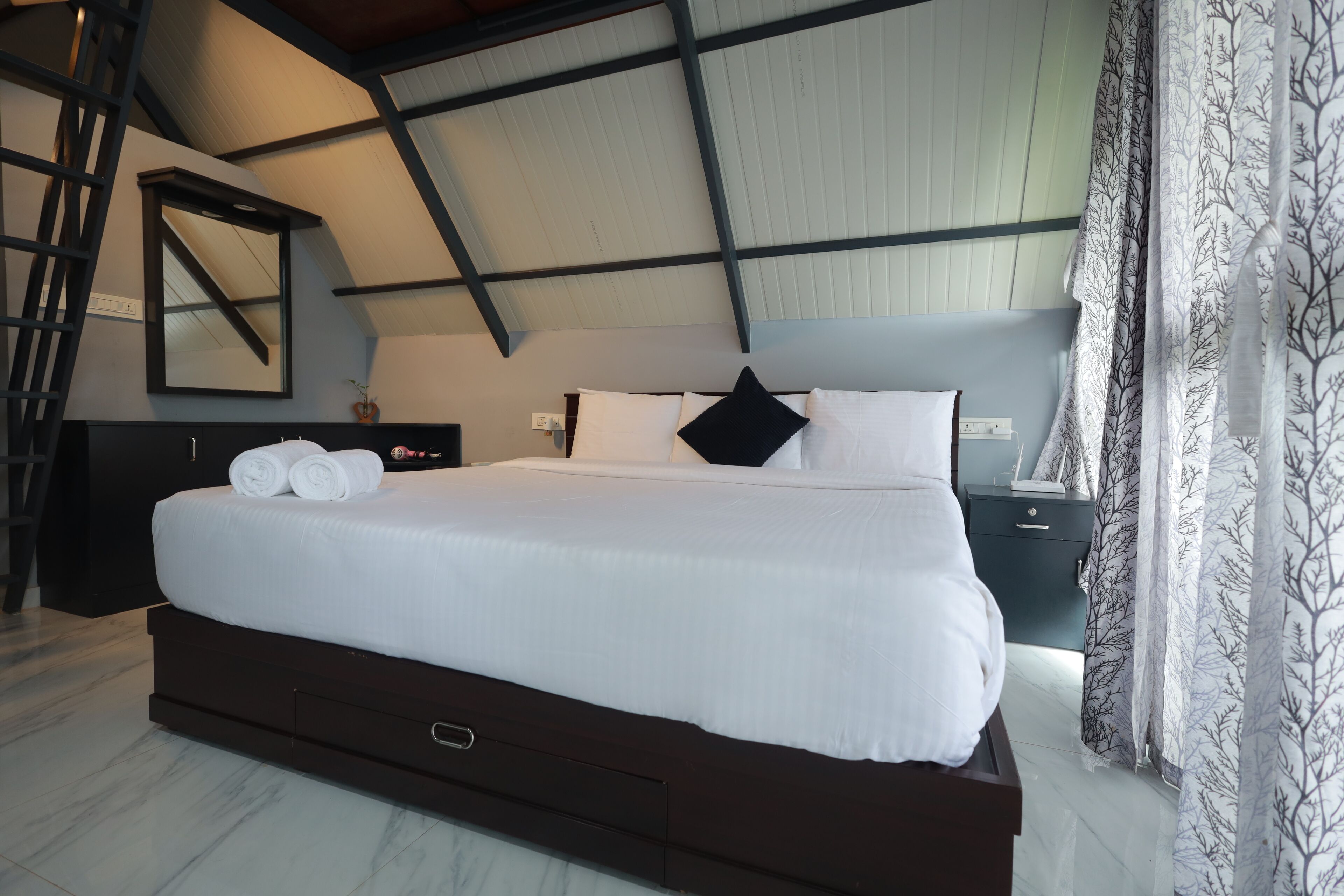 Una cama grande con ropa de cama blanca y un almohadón negro, un aparador de madera oscura, una mesita de noche con una lámpara y una escalera apoyada en la pared.