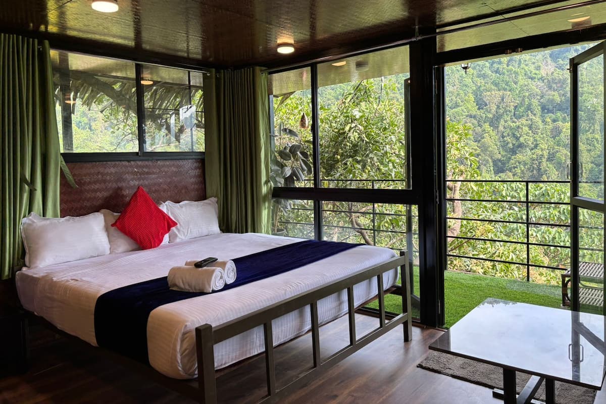 Quarto com cama grande, mesa pequena e vista para uma floresta exuberante através de grandes janelas.