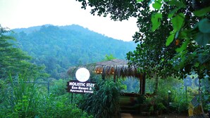 A sign for Holistic Stay Eco-Resort & Ayurvedic Retreat surrounded by lush greenery and a thatched-roof structure. - Holistic Stay Eco Resort (Talipparamba)