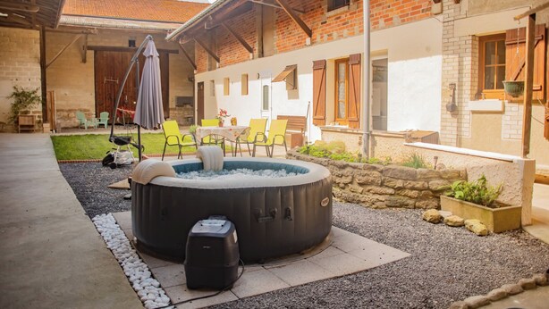 A hot tub in a courtyard with chairs and a table, surrounded by a brick building with a tiled roof. - Le Paradis des Bulles (Lisse-en-Champagne)