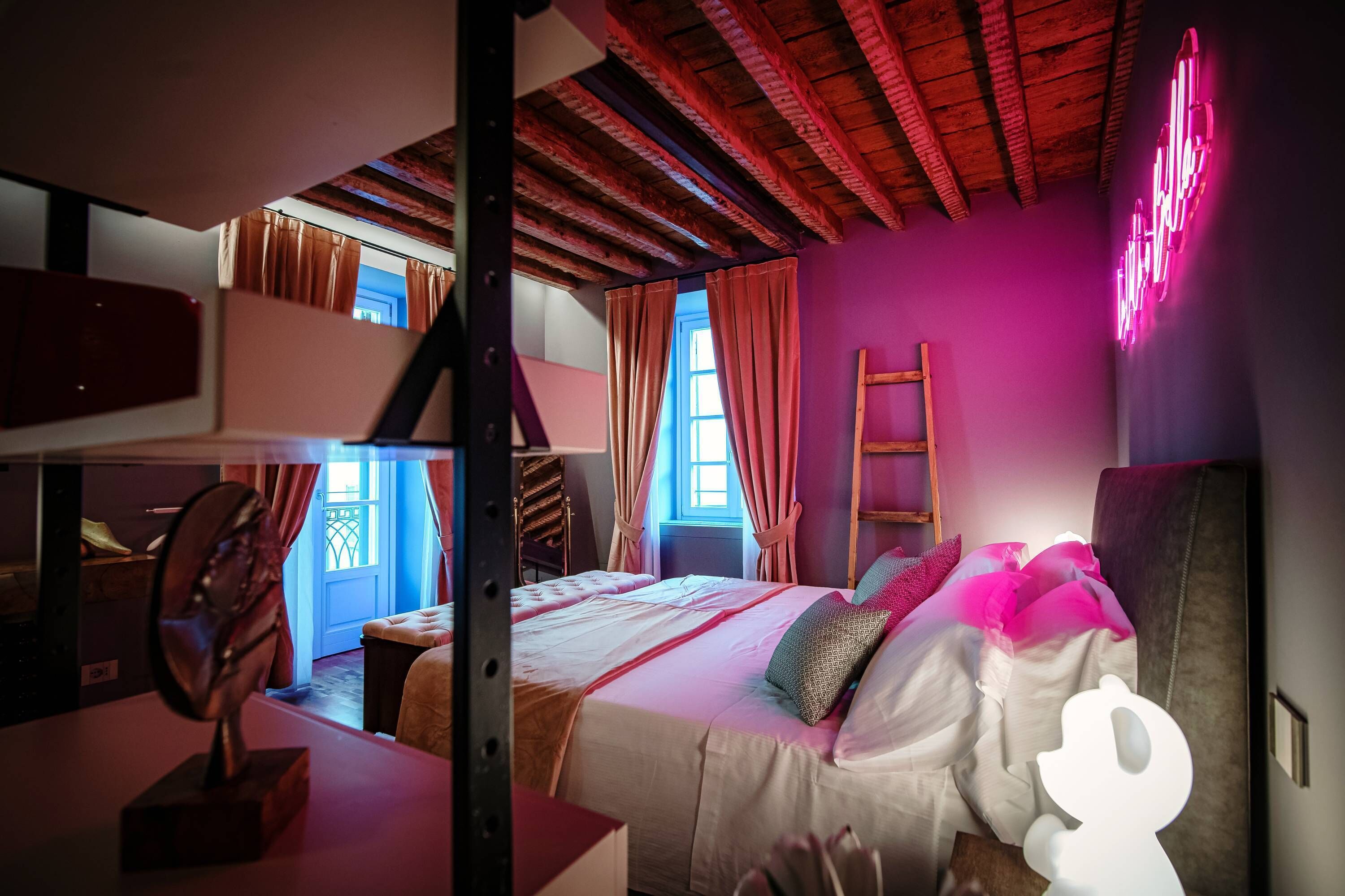 A bunk bed room with a neon sign, wooden beams, and a window with curtains.