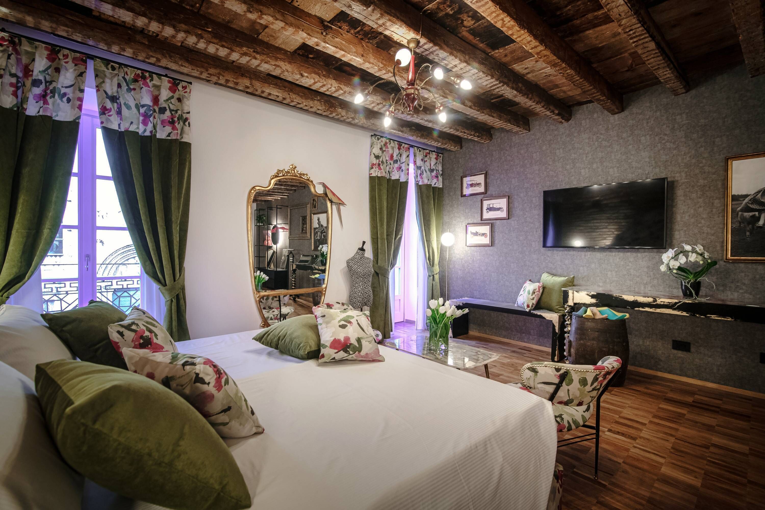 A bedroom with a wooden ceiling, a bed with floral pillows, a mirror, a television, and a desk with a vase of flowers.