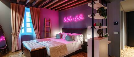 A bedroom with a bed, a vanity, a neon sign, and a window with curtains.