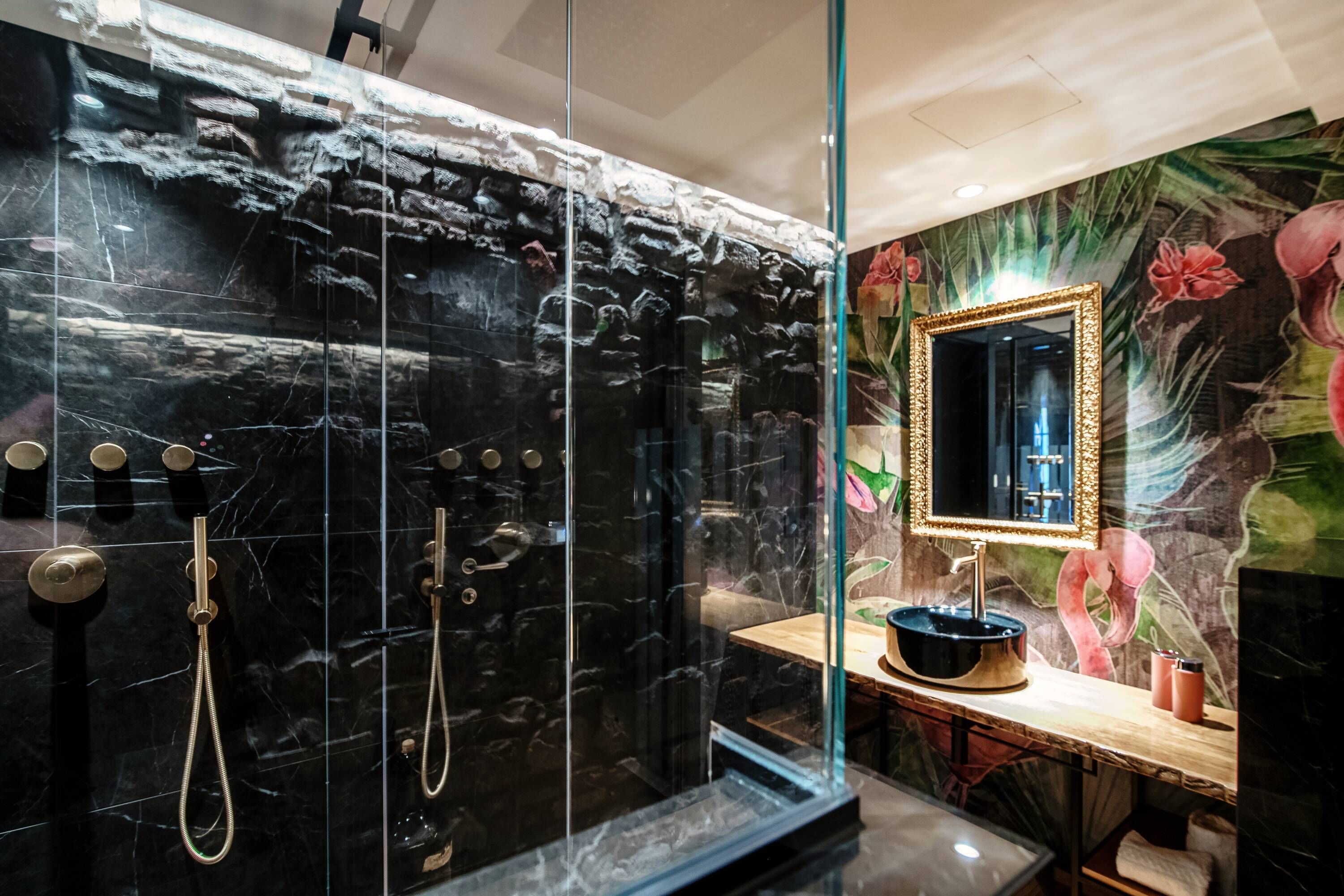 A modern bathroom with a black marble wall, a glass shower enclosure, a wooden vanity, and a mirror with a gold frame.