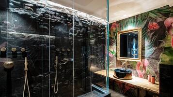 A modern bathroom with a black marble wall, a glass shower enclosure, a wooden vanity, and a mirror with a gold frame.