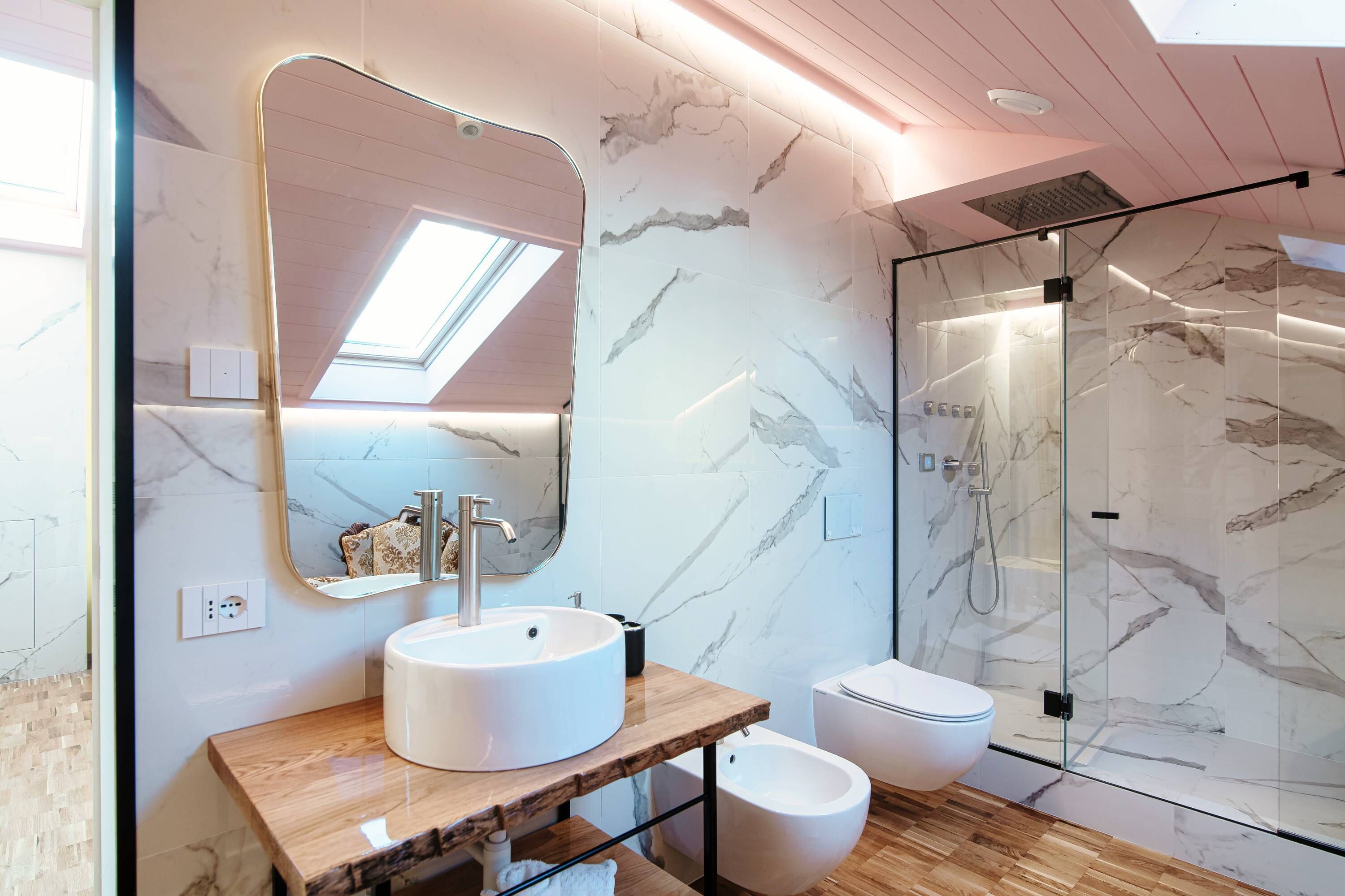 Ein modernes Badezimmer mit Oberlicht, Marmorwänden, einem Holzwaschtisch, einem weißen Waschbecken, einem Bidet und einer Duschkabine aus Glas.