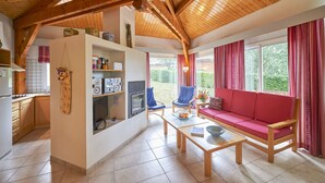A cozy living room with a red sofa, wooden coffee table, and blue chairs. A kitchen with a refrigerator and cabinets is visible in the background. - FranceComfort - L'Espinet (Quillan)