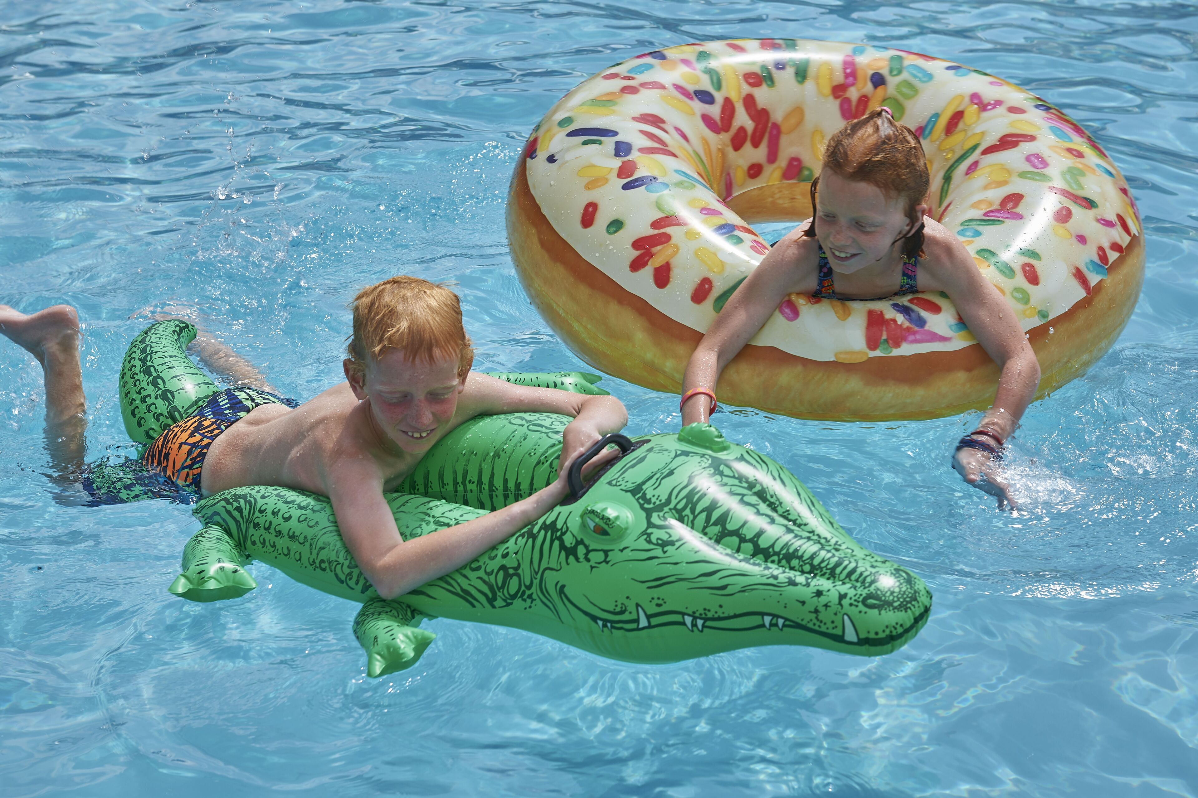 Dos niños flotando en una piscina con juguetes inflables, uno con forma de cocodrilo y el otro de rosquilla.