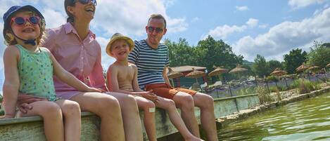 Eine Familie sitzt auf einer Betonbank an einem Gewässer, im Hintergrund sind Sonnenschirme und Grünpflanzen zu sehen.