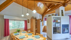 A dining area with a wooden table set for four, a kitchen in the background, and a fireplace with a TV. - FranceComfort - L'Espinet (Quillan)