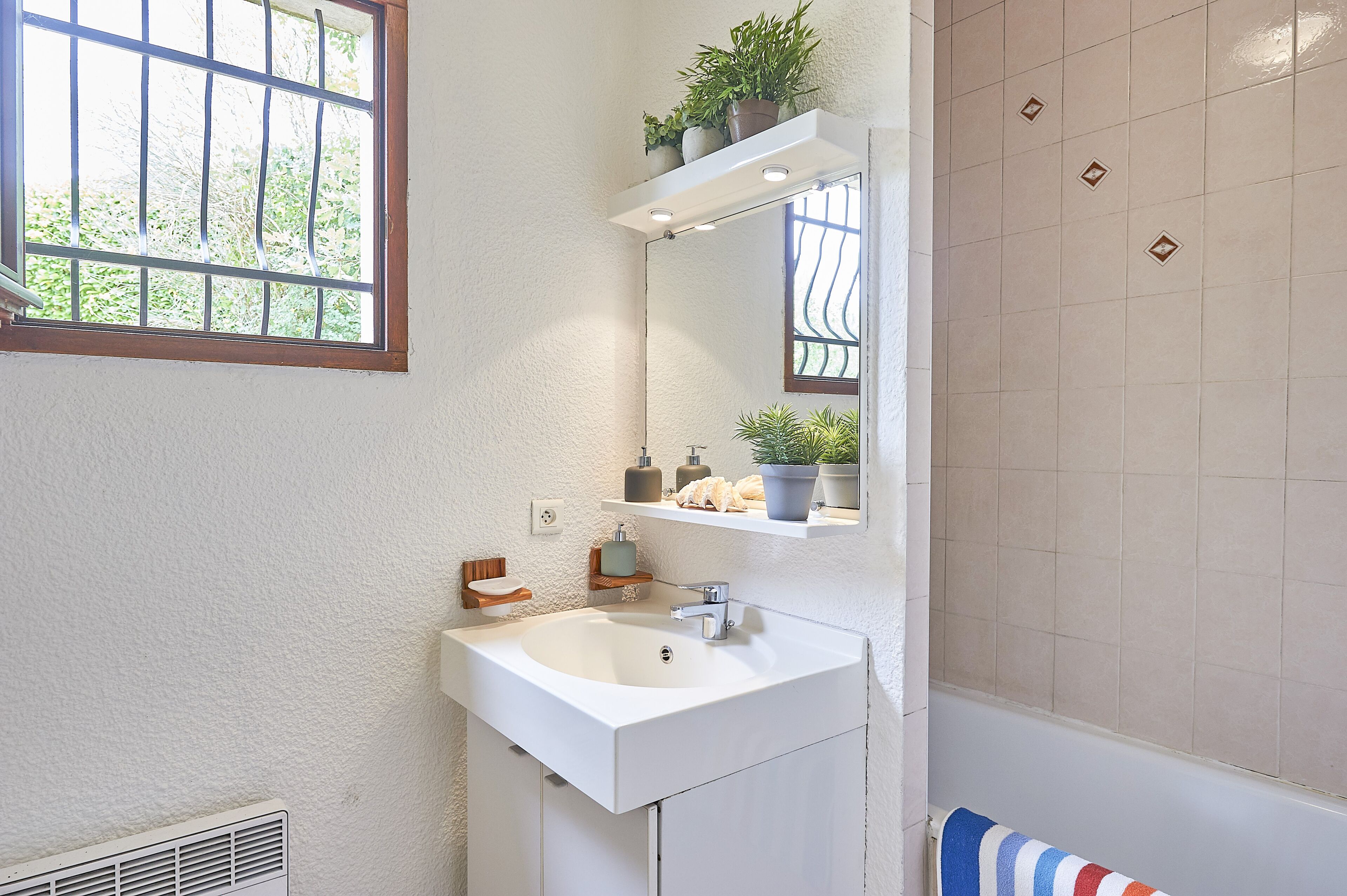 Un baño con lavamanos blanco, un espejo y una ventana con barrotes. Se observan estantes con artículos de aseo y un perchero para toallas.