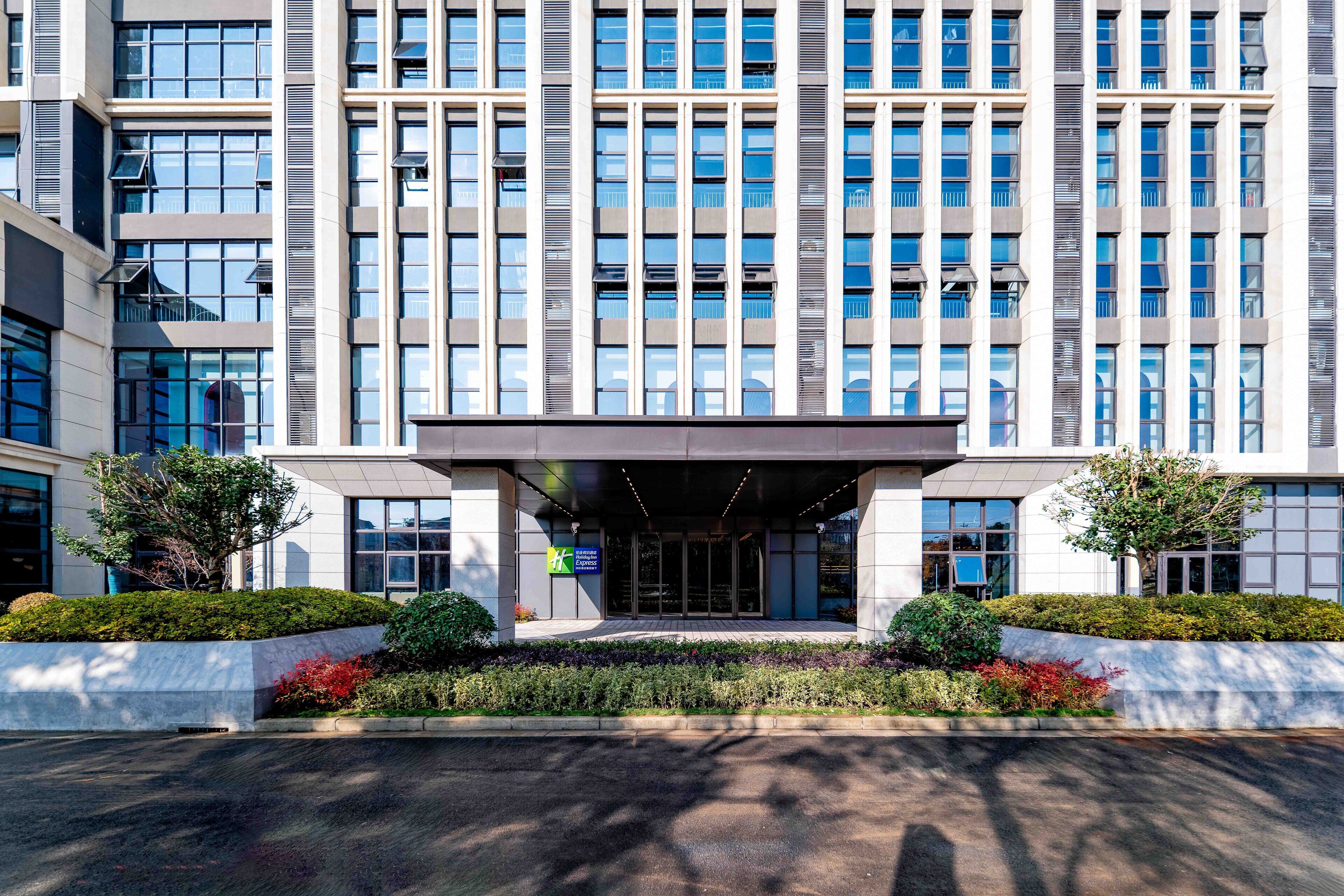 A modern building with a large entrance, multiple windows, and a landscaped area with shrubs and trees.
