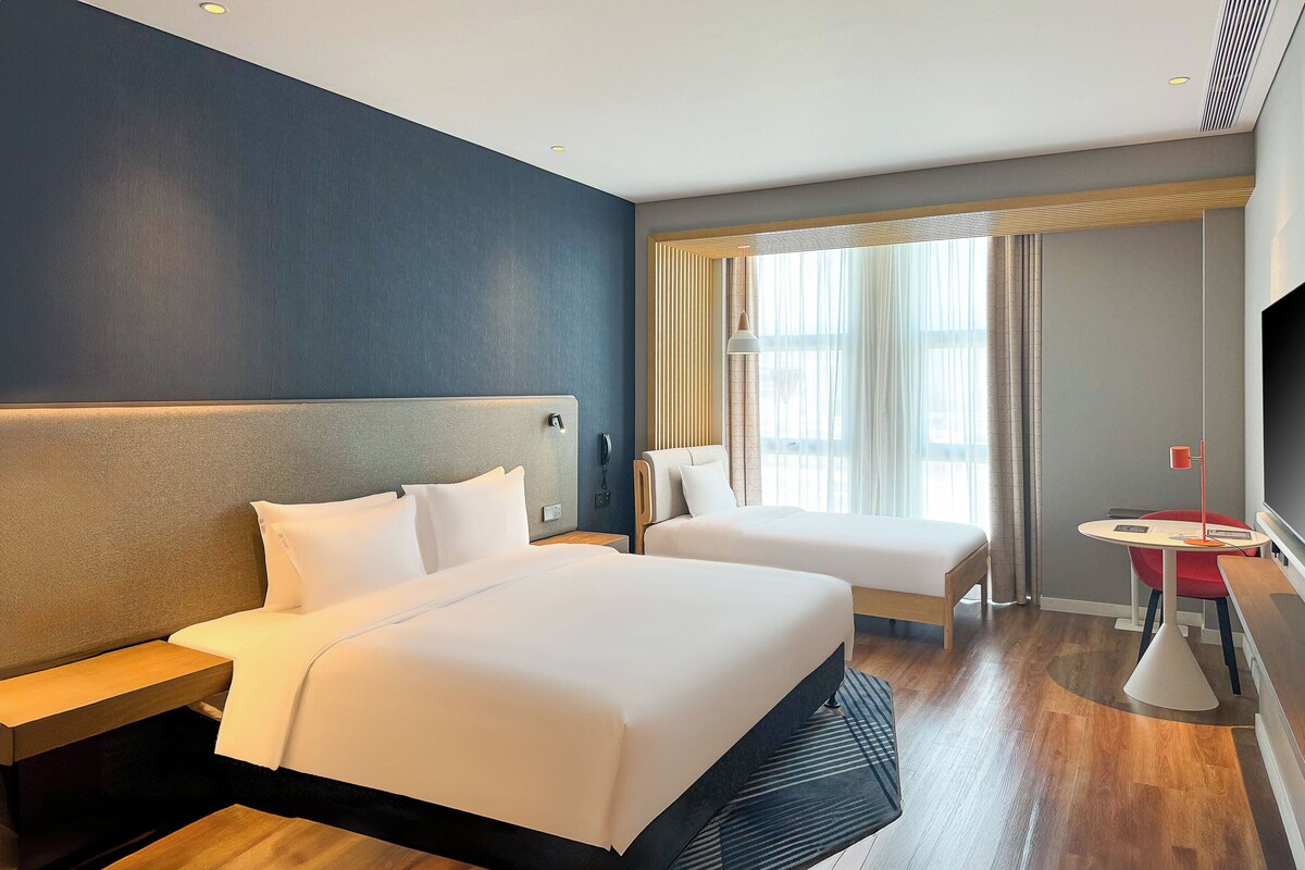A hotel room with two beds, a small table, a TV, and a window with curtains.