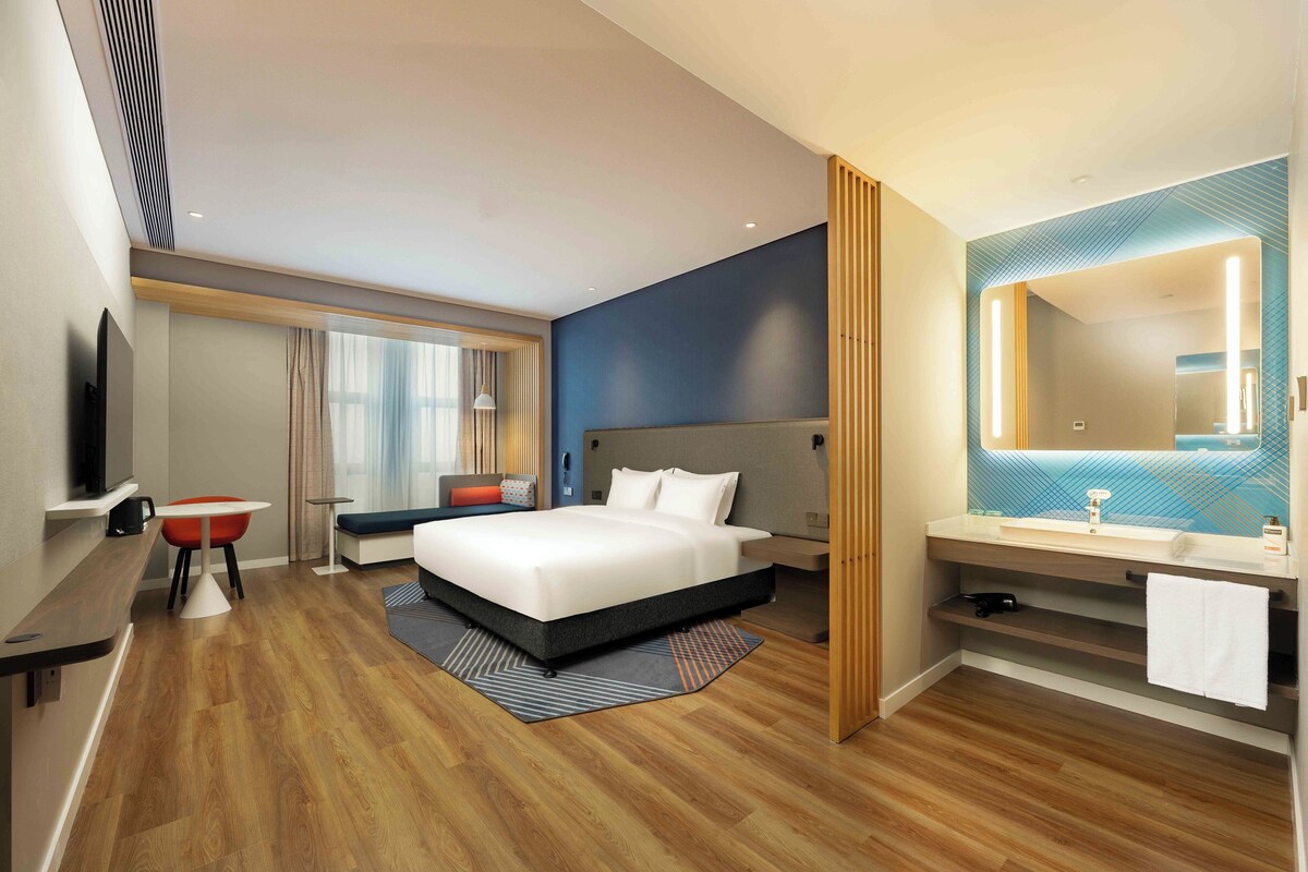 A modern hotel room with a large bed, a desk, a chair, a TV, a bathroom with a mirror and sink.