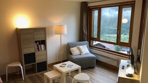 Una habitación acogedora con sofá, una mesita, un escritorio con lámpara, estantería y ventana con vistas a la vegetación.