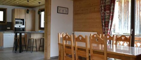 A wooden interior with a dining table and chairs, a kitchen area with a microwave, and a window with curtains.