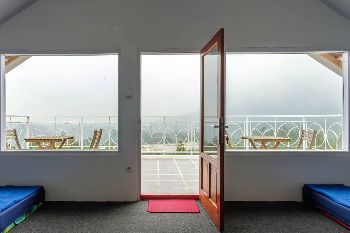 Ein Zimmer mit einer großen offenen Tür, die auf einen Balkon mit Blick auf einen Parkplatz und einen Zaun führt. Auf dem Balkon befinden sich zwei blaue Bänke und ein Holztisch.