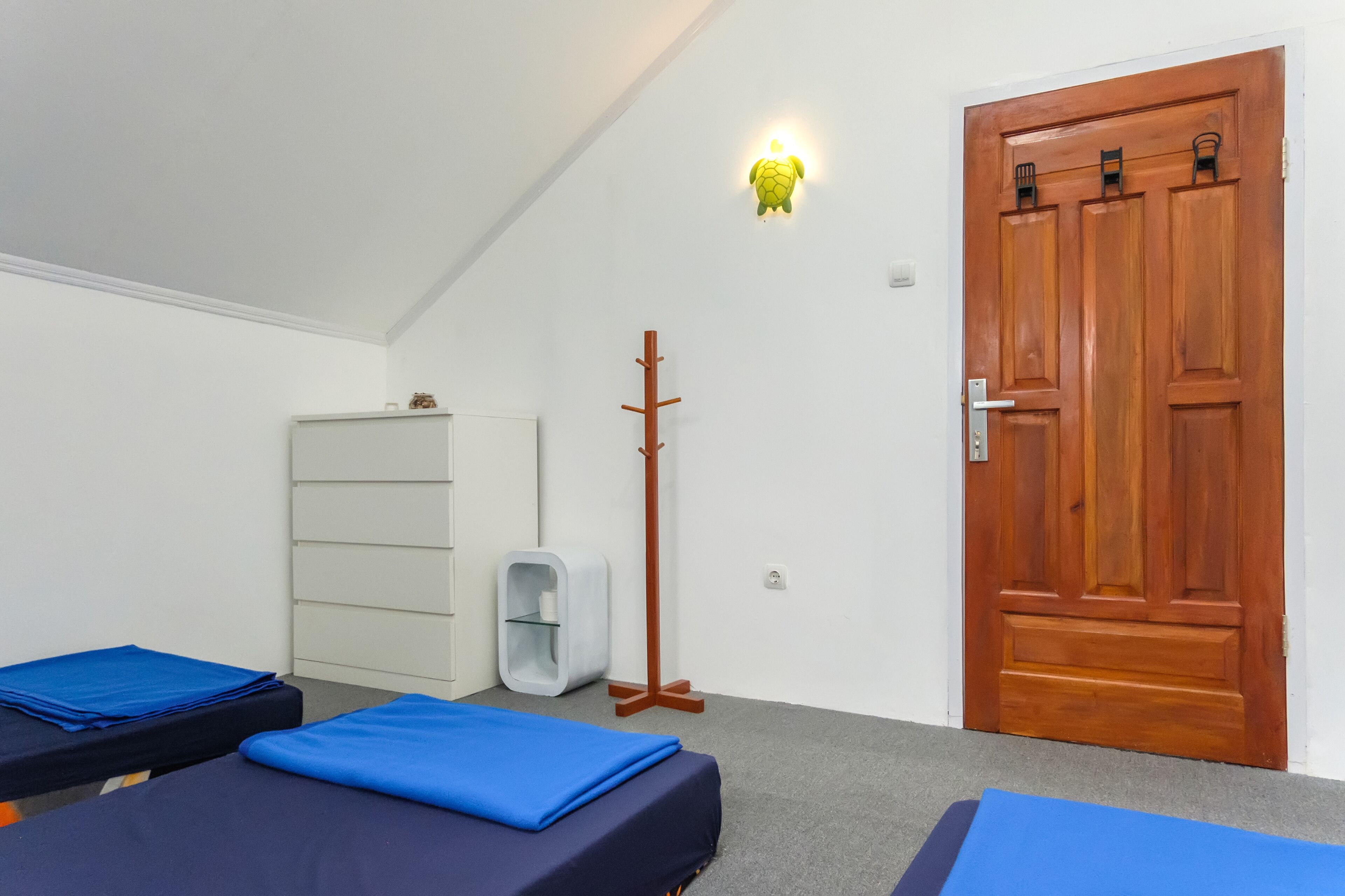 A room with two beds, a wooden door, a white wall, a shelf, and a wall-mounted light.