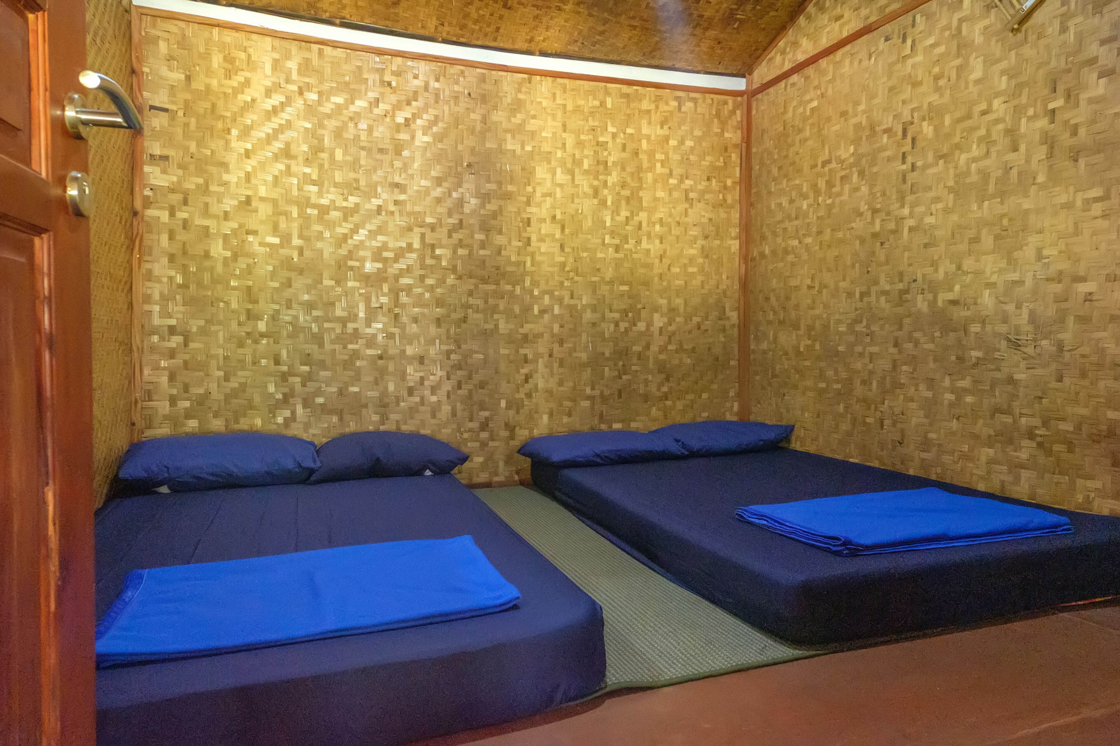 Habitación con dos camas, cada una con ropa de cama y almohadas azules, y una pared de bambú trenzado.
