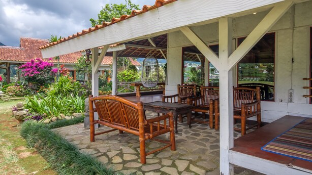 Un espacio al aire libre cubierto con mobiliario de madera y piso de piedra, rodeado de exuberante vegetación y un jardín.