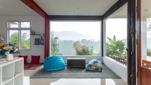A modern room with a large window, a blue bean bag, a small wooden chest, and a white shelf with decorative items. - Hilltop Camp, Lembang (Lembang)
