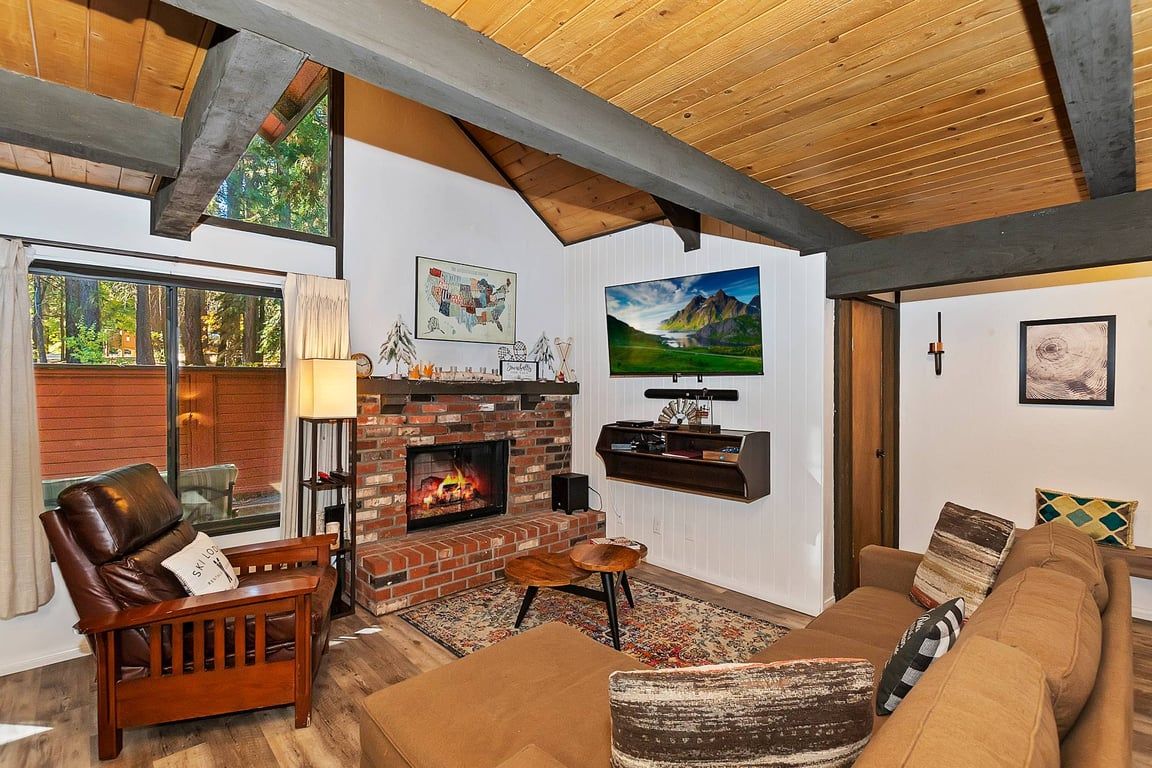 A cozy living room with a fireplace, wooden ceiling beams, a large window, and a mounted television.