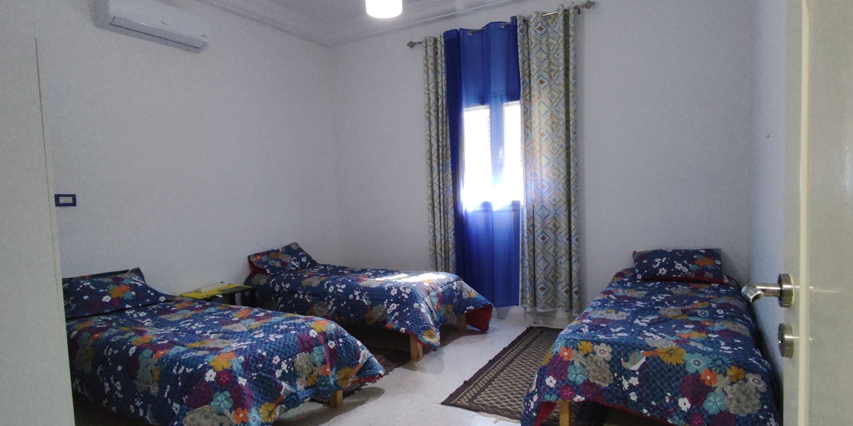 A room with two beds, a window with blue curtains, and a door.