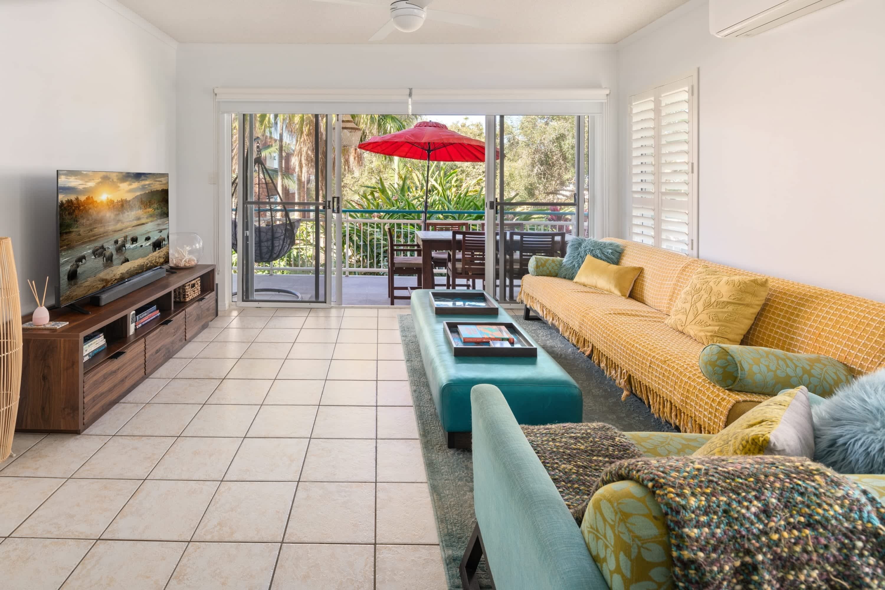A living room with a TV, sofa, ottoman, and a balcony with a red umbrella.