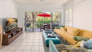 A living room with a TV, sofa, ottoman, and a balcony with a red umbrella. - Sunny Seaside Retreat in Coolum Beach (Coolum Region)