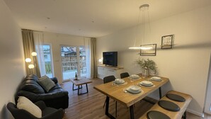 Ein modernes Wohnzimmer mit einem Holzes Esstisch, gedeckt für vier Personen, einer Couch und einem Fernseher.